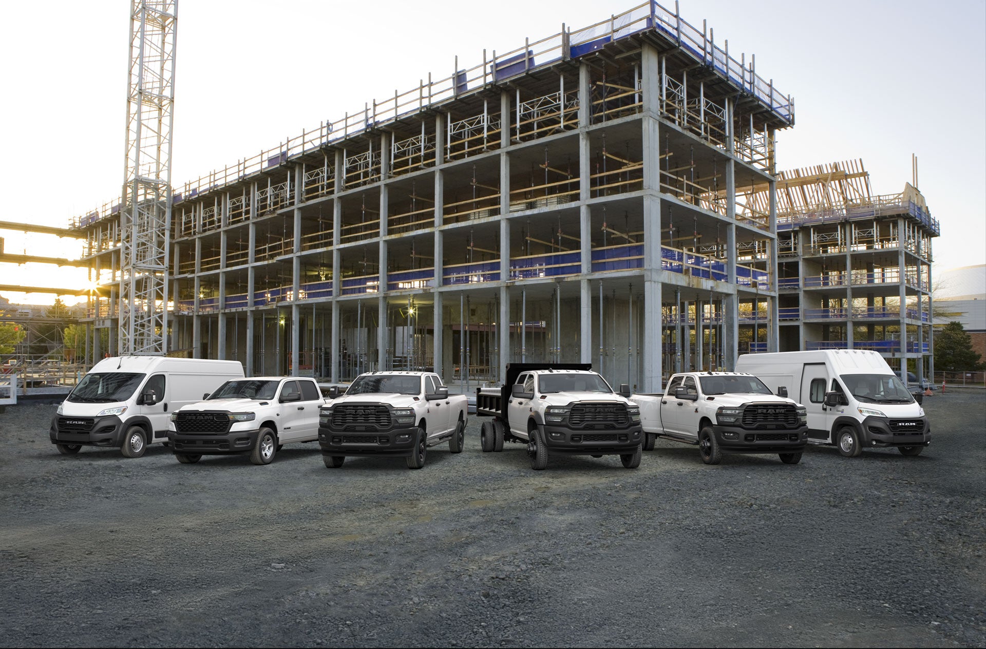 Una flota de camionetas y furgonetas Ram blancas aparcada en un terreno de grava frente a un edificio de hormigón en construcción.